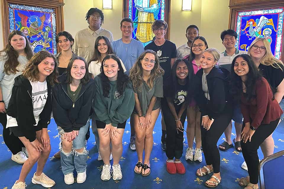 A group of 17 SLU students pose for a photo in a room at DuBourg Hall with stained glass windows behind them.