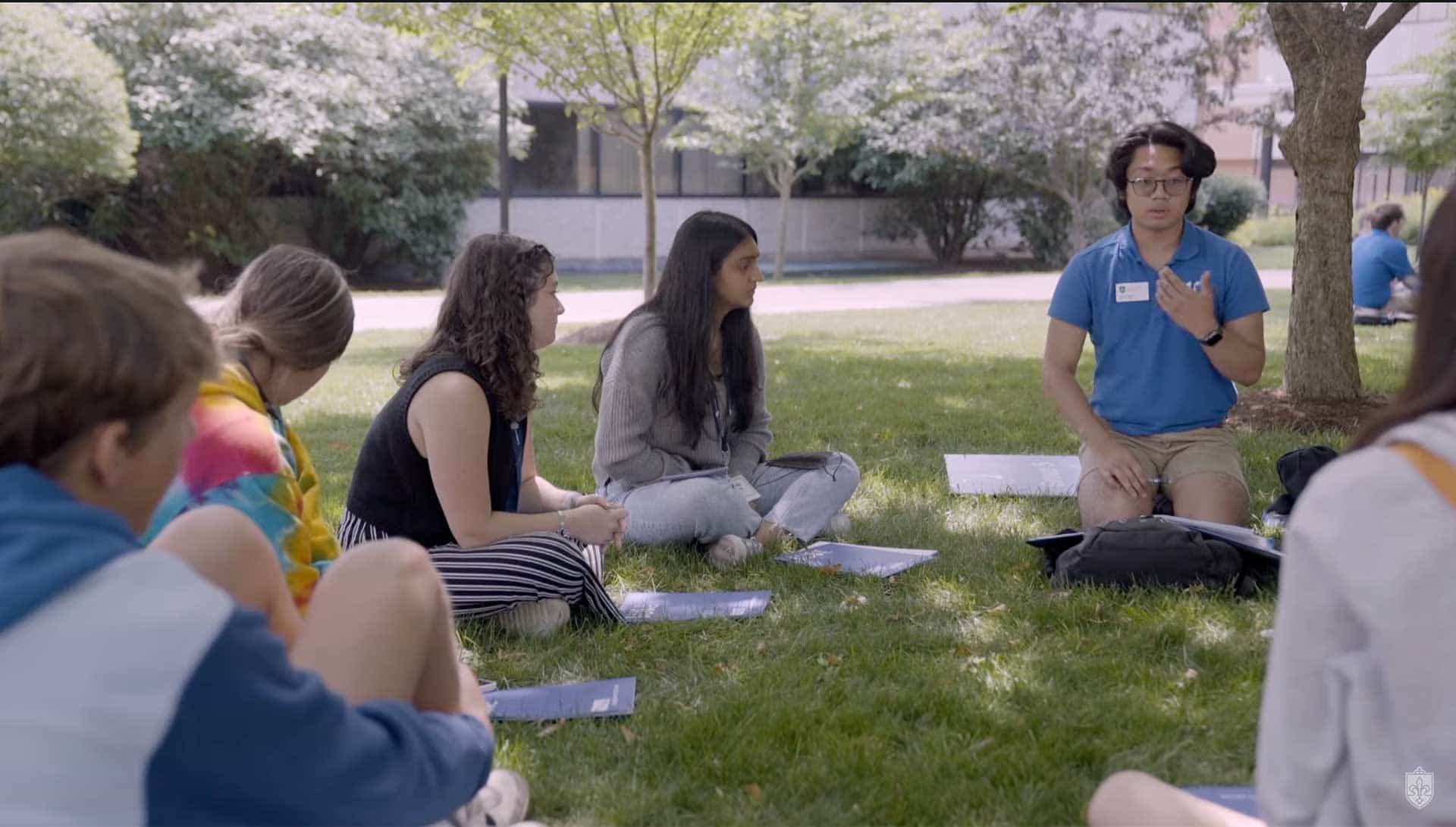 A SLU 101 leader speaks to a group of incoming students. The group is sitting in a circle on the ground under a tree near Ritter Hall.