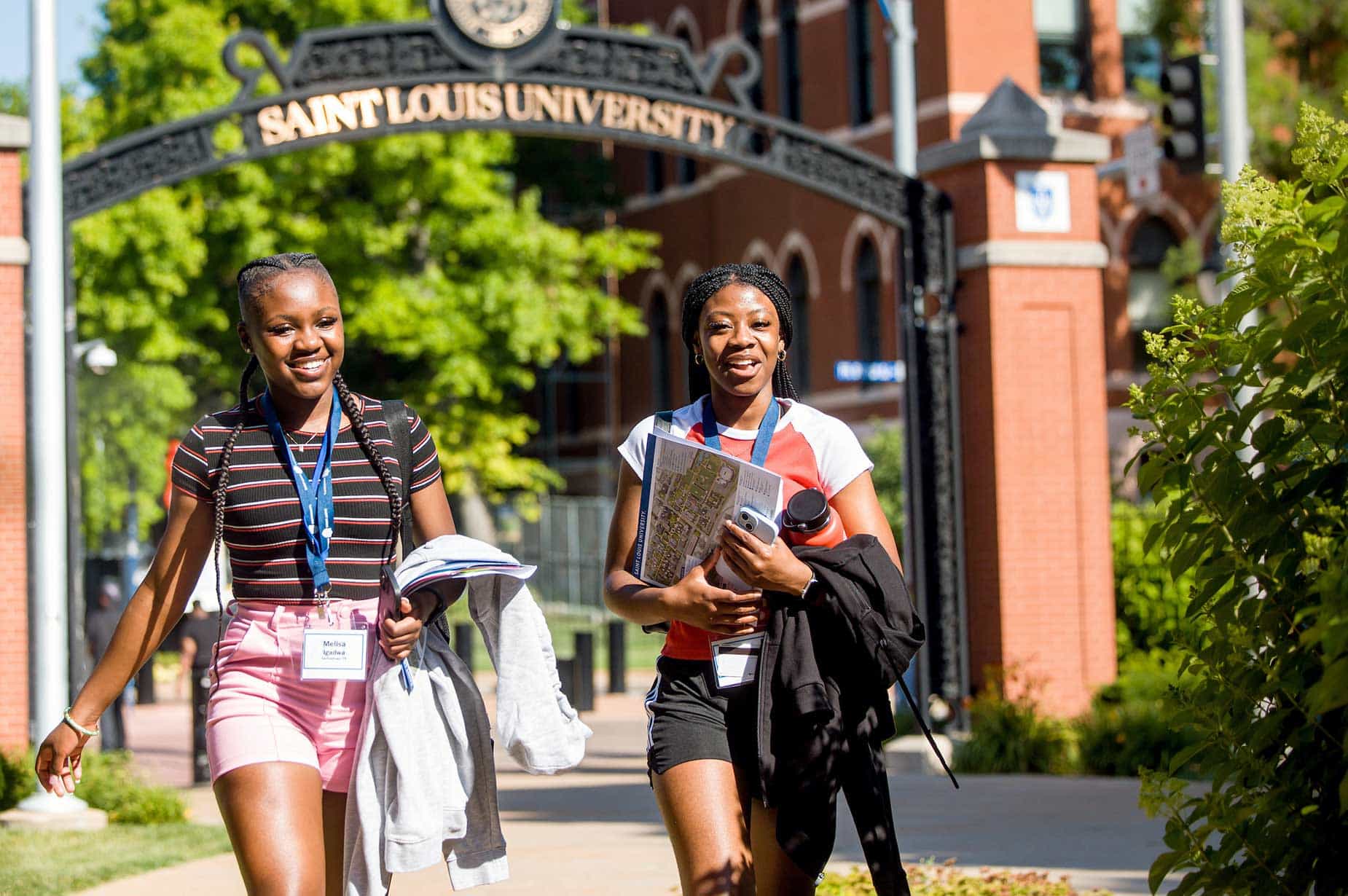 Two female students, wearing SLU 101 lanyards and holding SLU 101 folders, walk along West Pine Boulevard on SLU’s campus on a sunny day.