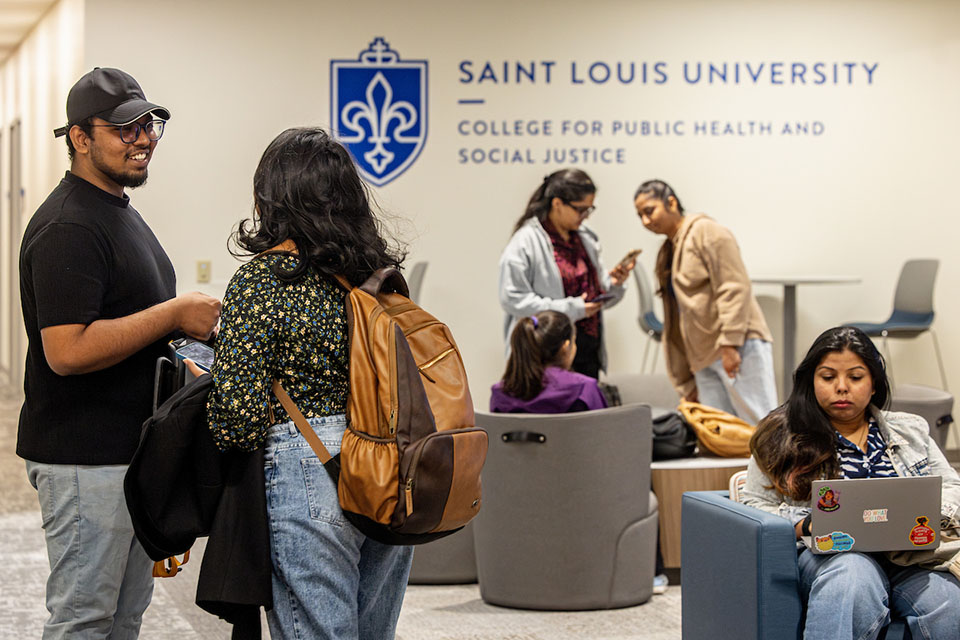 Students gather in a room on campus. The wall has a sign that says Saint Louis University College for Public Health and Social Justice.