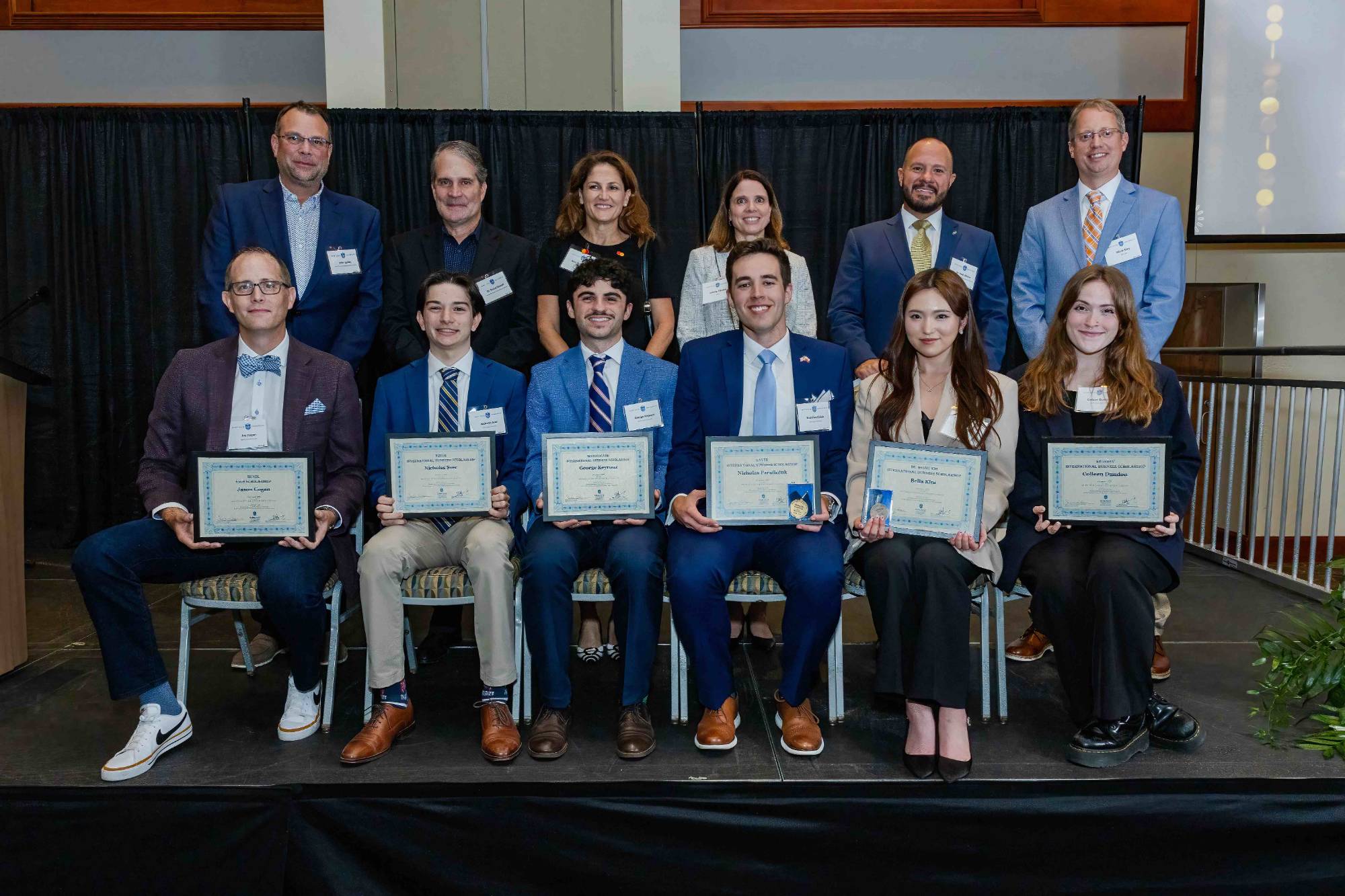 2025 Award Winners Award winners sit in a row holding up awards plaques while faculty stand behind them.