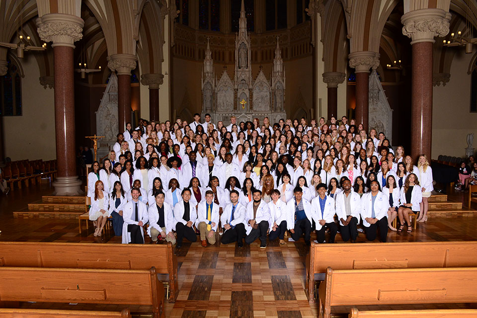 SLU's Trudy Busch Valentine School of Nursing students celebrate receiving their white coats. A group of students wearing white coats pose for a photo inside a dimly lit church.