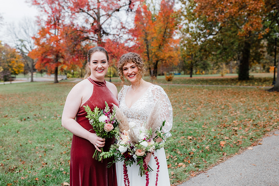 Two women dressed in wedding attire pose for a photo outdoors on a fall day.