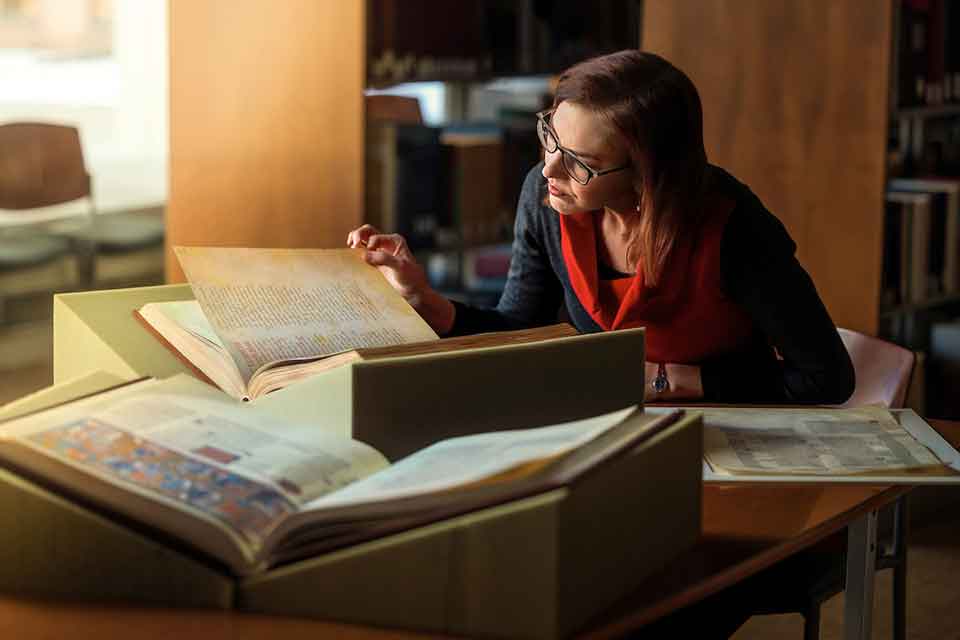 A woman wearing glasses sits at a table in a library, examining large open books with special mounts.